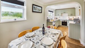 A dining area with a table set for four, a kitchen in the background, and a window with a view of greenery.