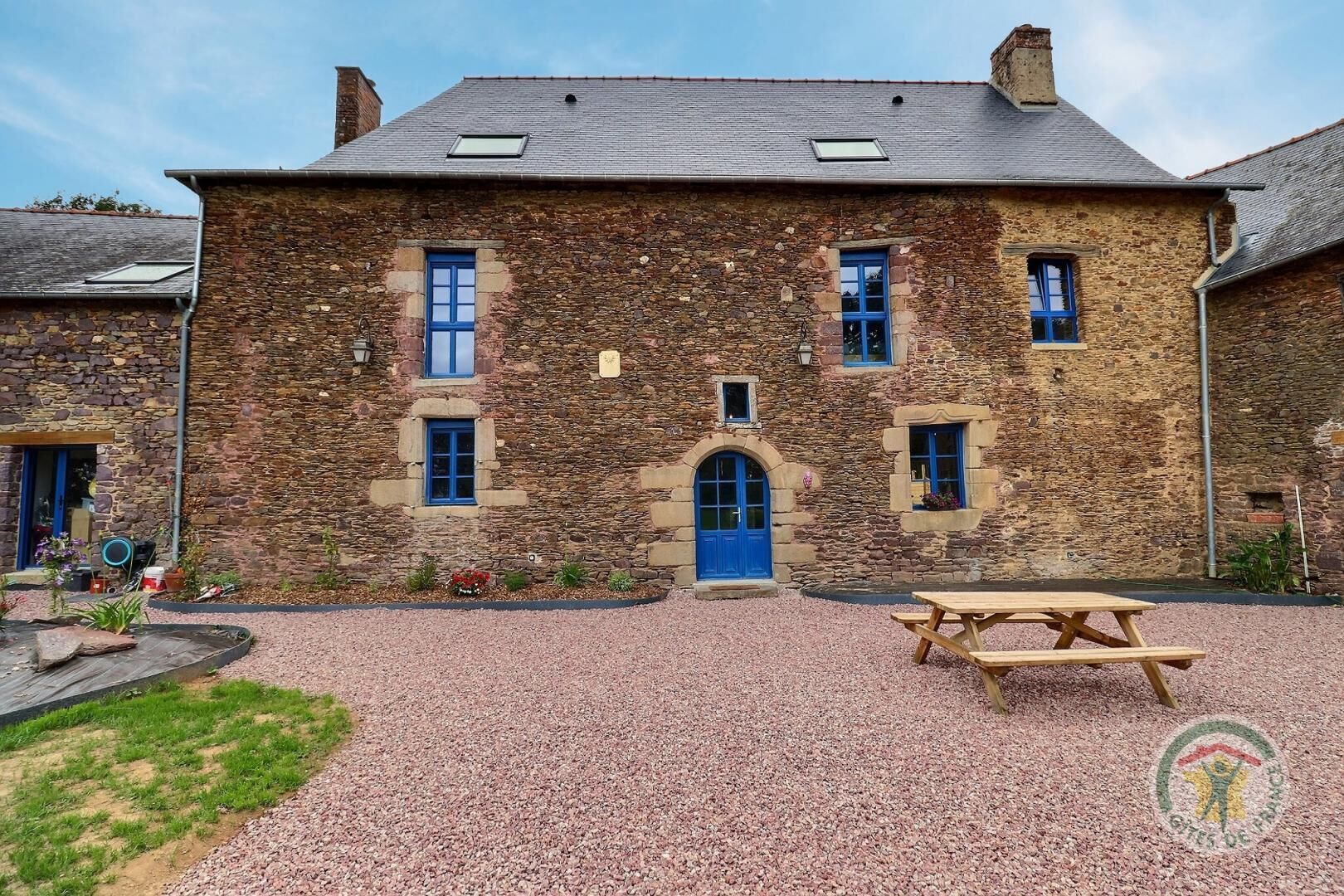 A stone building with a central arched entrance and multiple windows, a gravel driveway, and a picnic table.