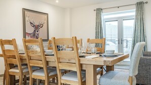 A dining area with a wooden table set for six, six wooden chairs with grey cushions, a framed deer painting on the wall, and a view of the city through a window with curtains. - Copper Beech Cottage - UK35892 (Ludchurch, near Narberth)