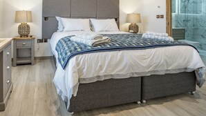 A modern bedroom with a large bed, two bedside tables, a nightstand, a wooden dresser, and a bathroom with a glass shower. - Copper Beech Cottage - UK35892 (Ludchurch, near Narberth)