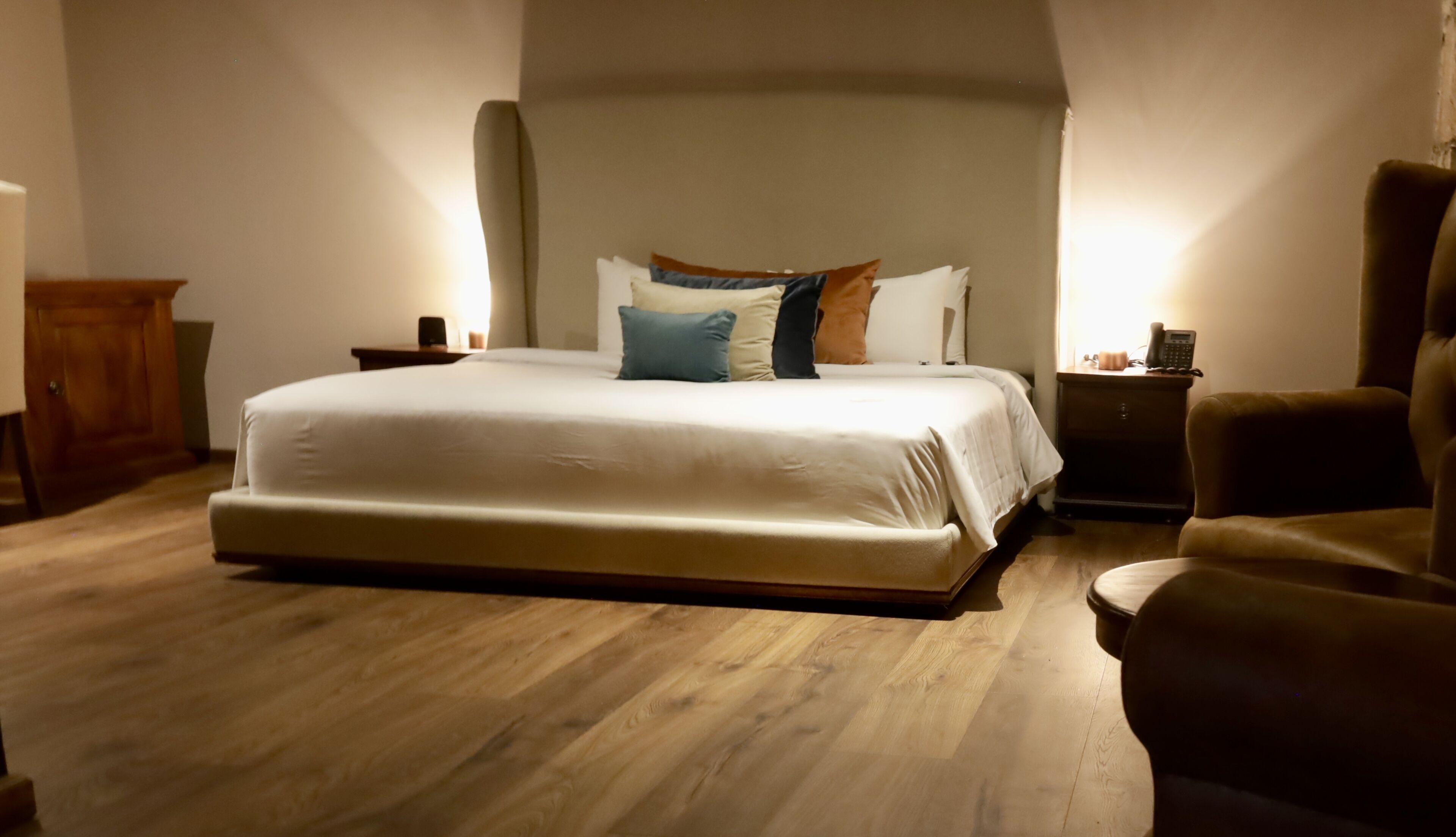 A bedroom with a large bed, a wooden floor, a nightstand with a lamp, and a chair.