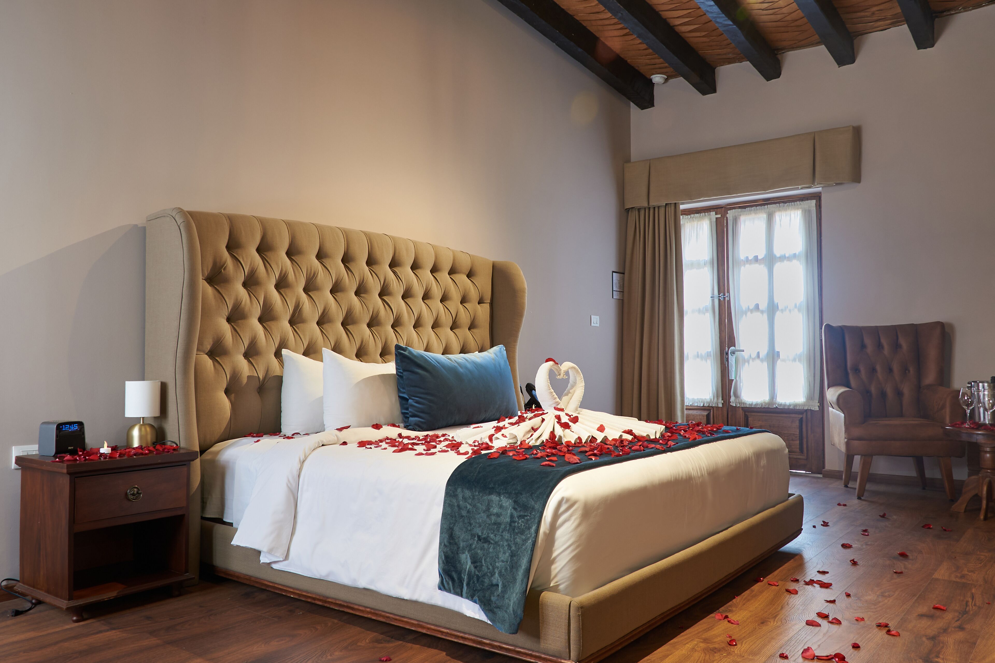 A large bed with a tufted headboard, a bedside table with a lamp, and a chair in a room with wooden flooring and a large window.