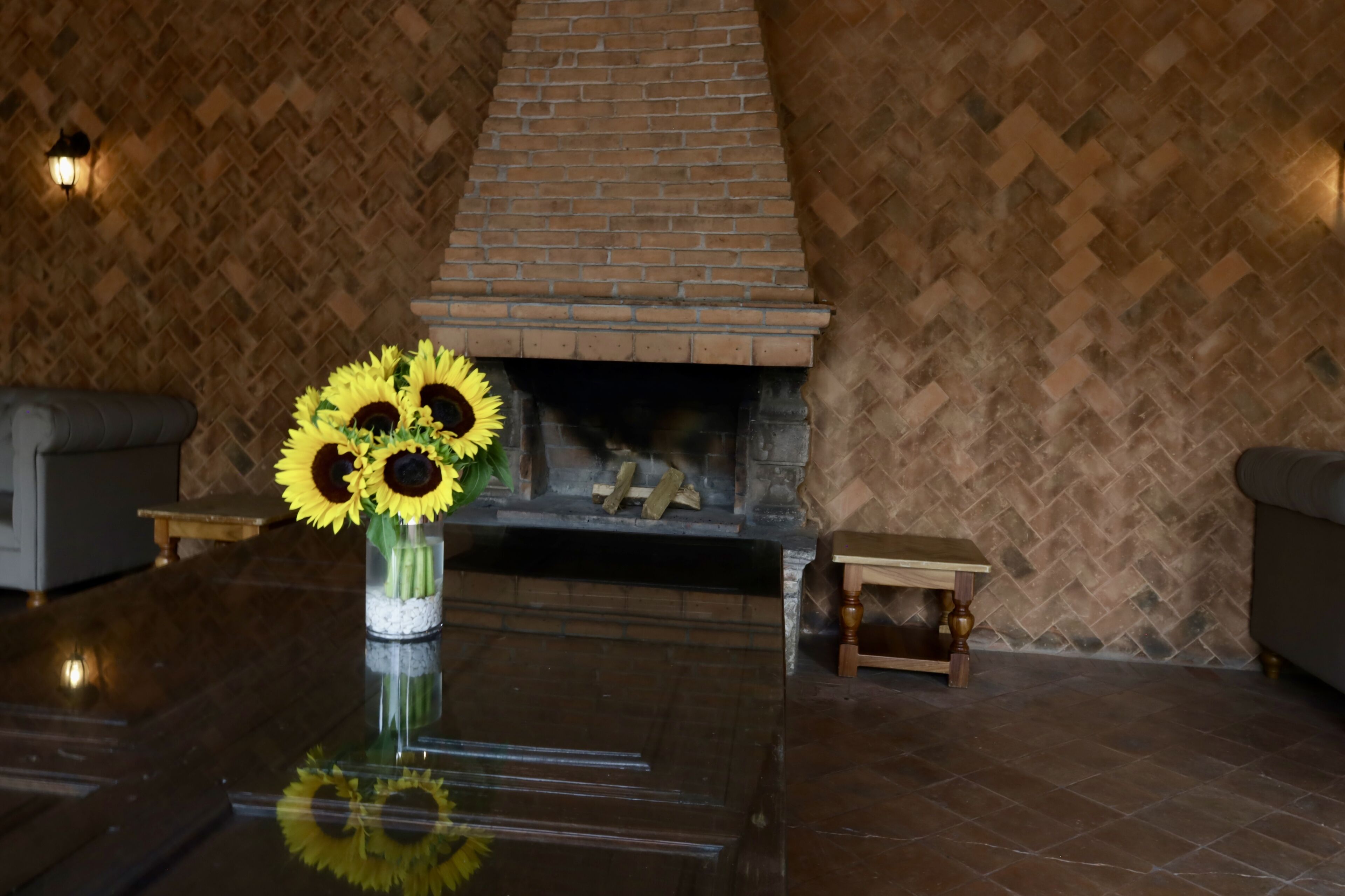 Una habitación con chimenea de ladrillo, un jarrón con girasoles y una pequeña mesa de madera.
