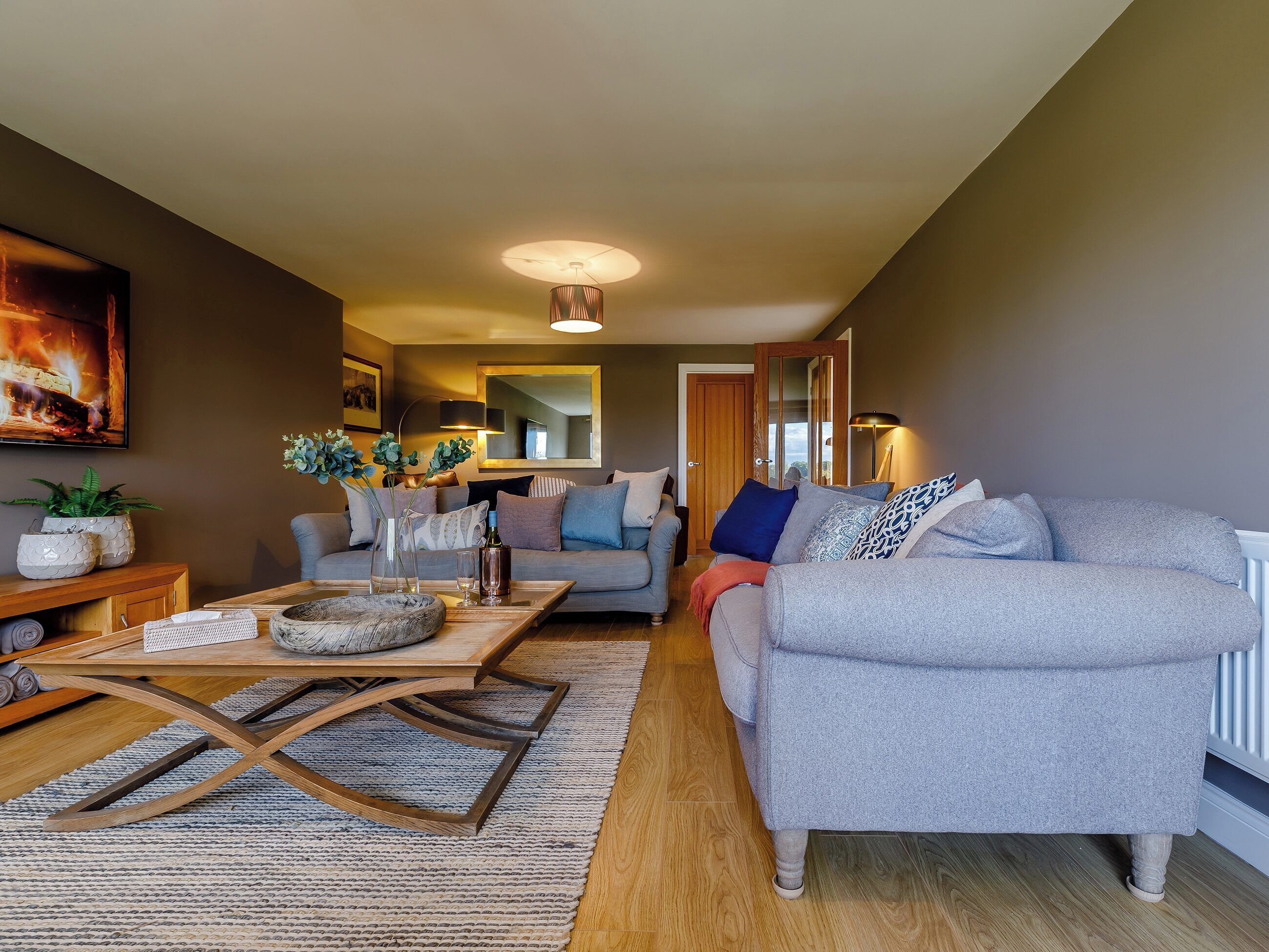 Un salon moderne avec un canapé gris, une table basse en bois et un téléviseur placé à gauche.