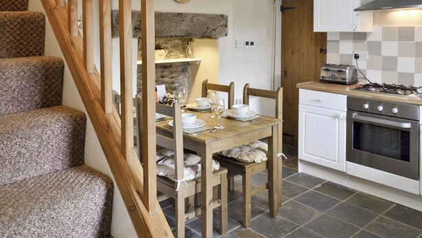 A kitchen with a wooden dining table set for two, a stove, and a clock on the wall. - Close to the village centre with many walks and cycle routes from the doorstep. (Bradwell)