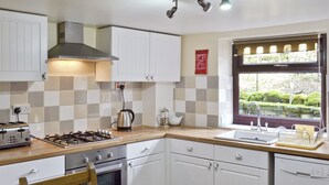A kitchen with white cabinets, a gas stove, a kettle, and a window with a view of greenery. - Close to the village centre with many walks and cycle routes from the doorstep. (Bradwell)