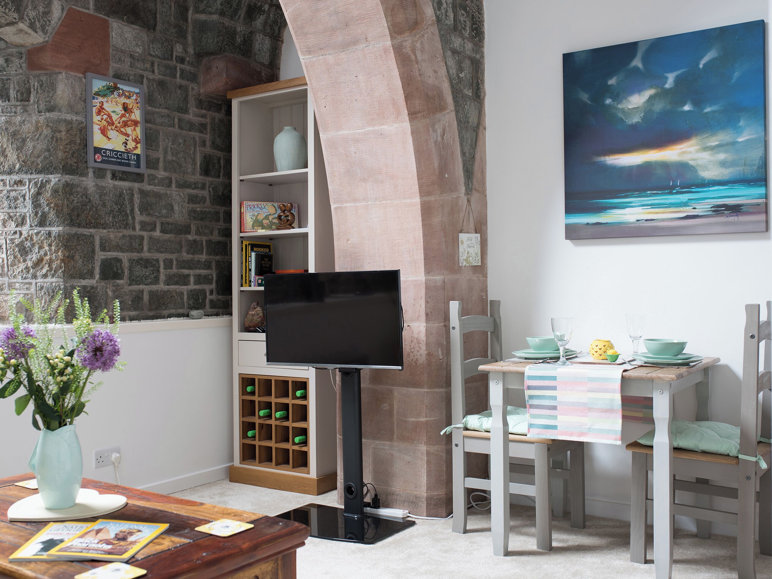 Habitació amb paret de pedra, televisió, taula de dinar amb cadires i una prestatgeria.