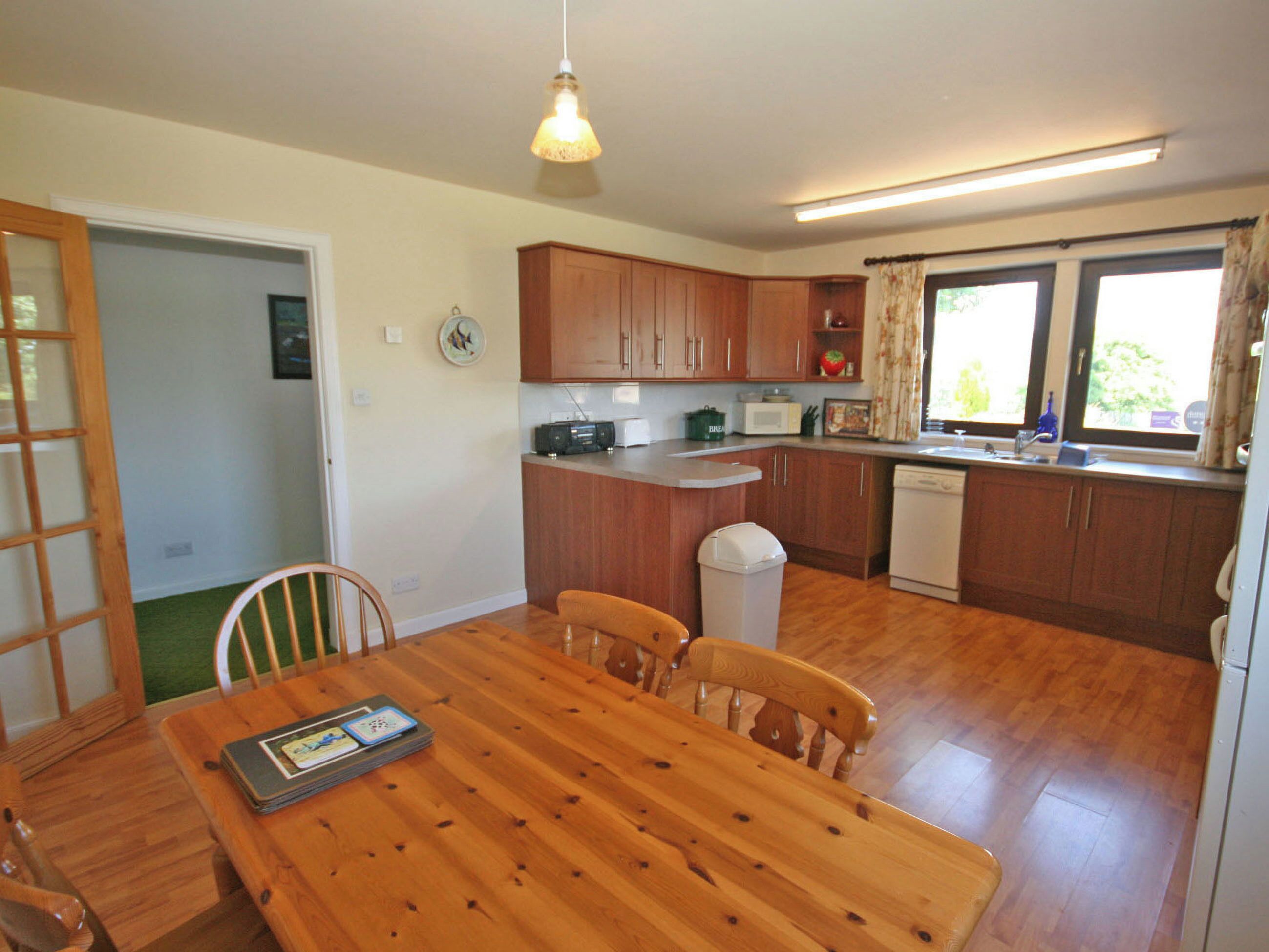 A compact kitchen with wooden cabinets, a dining table with chairs, a trash bin, and a window with curtains.