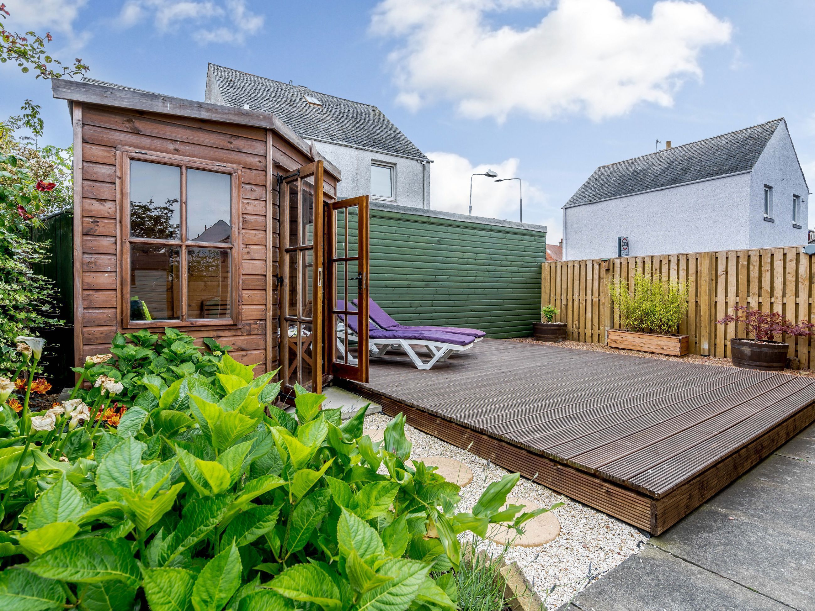 A wooden cabin with a deck, a green wall, and a white house in the background.