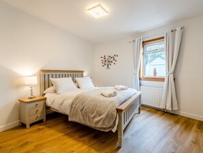 A bedroom with a wooden bed, white bedding, a nightstand with a lamp, a radiator, and a window with curtains.