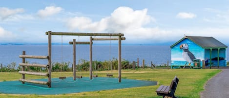 A playground with swings and a bench, a small building with a mural, and a view of the sea.