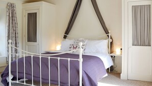 Un dormitorio con estructura de cama metálica blanca, ropa de cama morada, una puerta blanca y una ventana con cortinas.