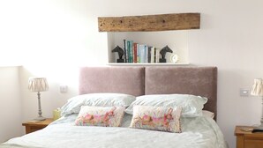 A bedroom with a bed, pillows, a nightstand, a lamp, a wooden shelf with books, and a clock.