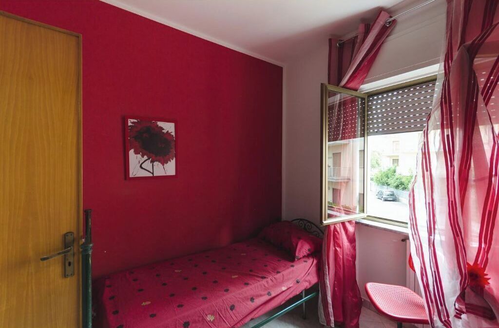 Una habitación pequeña con una cama individual, una ventana con cortinas, una puerta y un cuadro colgado en la pared.
