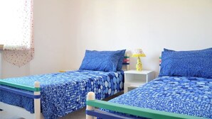 Ein Schlafzimmer mit zwei Betten, jeweils mit blauer Bettwäsche und Kissen, einem kleinen Nachttisch mit Lampe und einem Fenster mit durchscheinendem Vorhang.