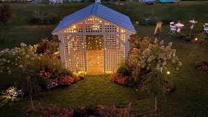 Un mirador con luces colgantes, rodeado de un jardín con diversas flores, cerca de un lago con barcos y casas al fondo.