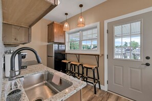 Cocina con encimera de granito, pileta de acero inoxidable, alacenas de madera y ventana con vista a árboles y edificios.
