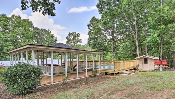 Een overdekte pergola met een houten terras, omgeven door bomen en een kleine schuur.