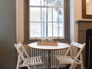 Une pièce avec un radiateur blanc, une table ronde sur laquelle se trouve un plateau rempli de tasses, et deux chaises blanches.