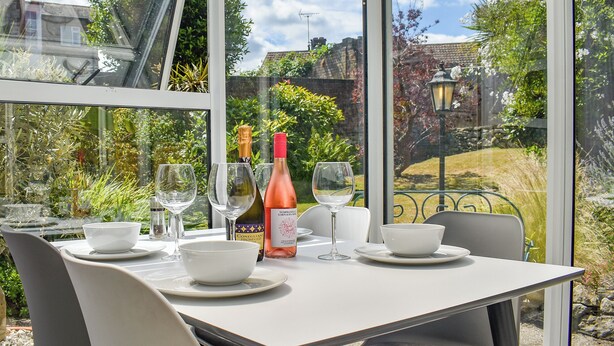 Mesa posta para dois, com taças de vinho, garrafa de vinho e tigelas, com vista para um jardim através de grandes janelas.