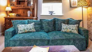 A teal-colored sofa with floral cushions, a small side table with a lamp, and a decorative bowl on a windowsill.
