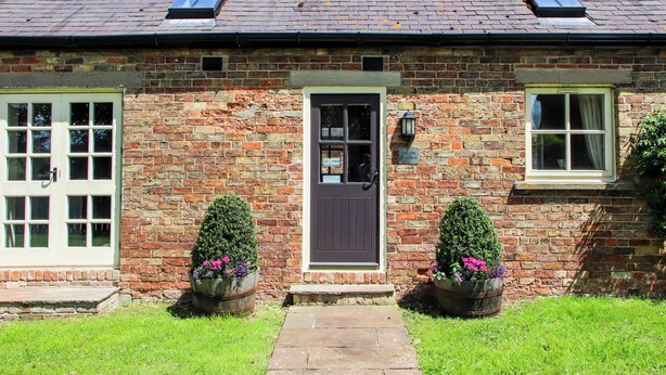 A brick building with a dark entrance door, two windows, and a small sign reading "The Old Cowshed."