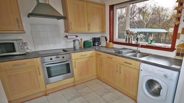 A kitchen with wooden cabinets, a microwave, an oven, a washing machine, and a window with a view of trees. - 2 bedroom accommodation in Portree, Isle of Skye (Portree, Isle of Skye)
