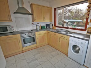 Een keuken met houten kastjes, een magnetron, een oven, een wasmachine en een raam met uitzicht op bomen.