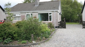 A two-story house with a tiled roof, a chimney, and a gravel driveway leading to a wooden gate. - 2 bedroom accommodation in Portree, Isle of Skye (Portree, Isle of Skye)