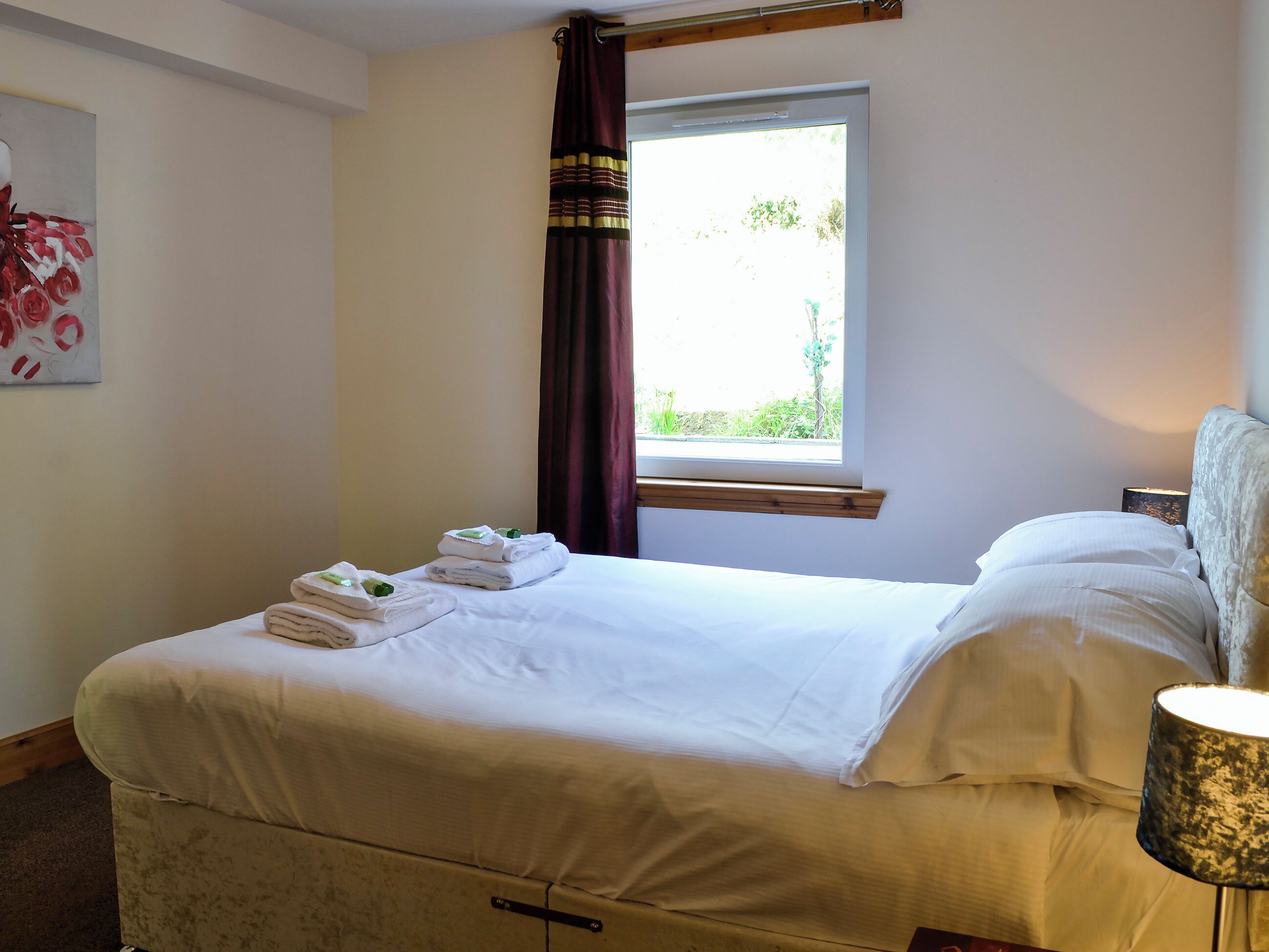 A hotel room with a bed, a window with curtains, a bedside lamp, and a painting on the wall.