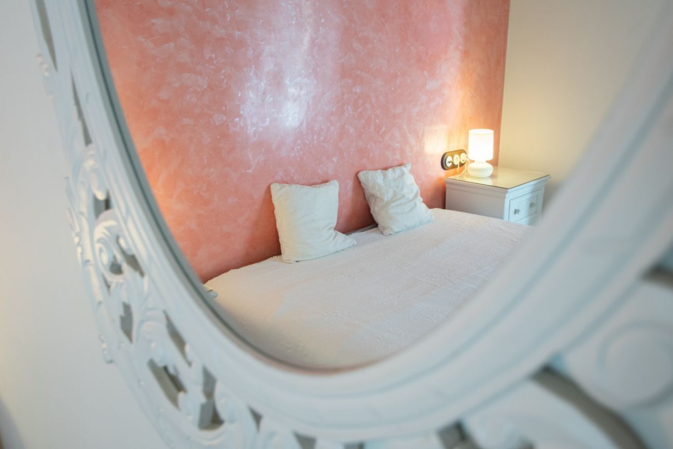 A bedroom with a pink wall, a white bed, two pillows, and a nightstand with a lamp.