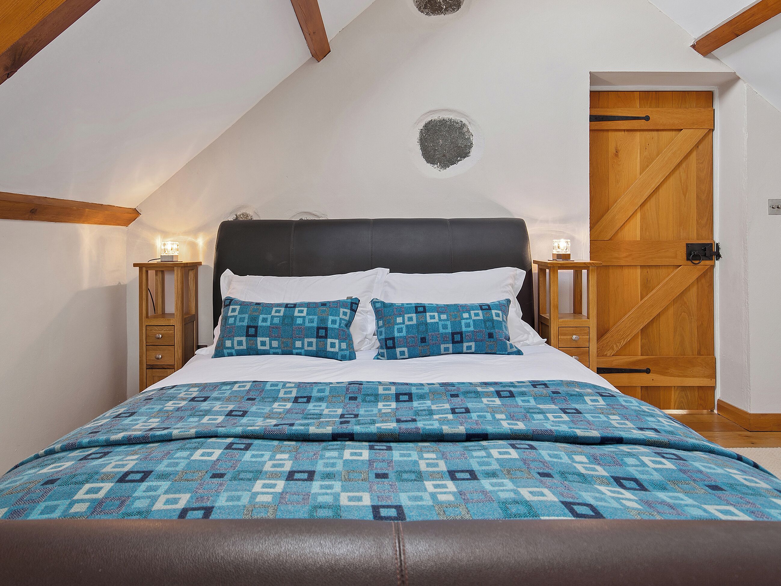 A bedroom with a bed, a wooden nightstand, a wall clock, a door, and a ceiling with wooden beams.