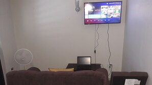 A room with a TV mounted on the wall displaying a streaming service interface, a laptop on a desk, a standing fan, and a chair with a document on it. - Cozy vacation apartment in Kingston (Kingston)