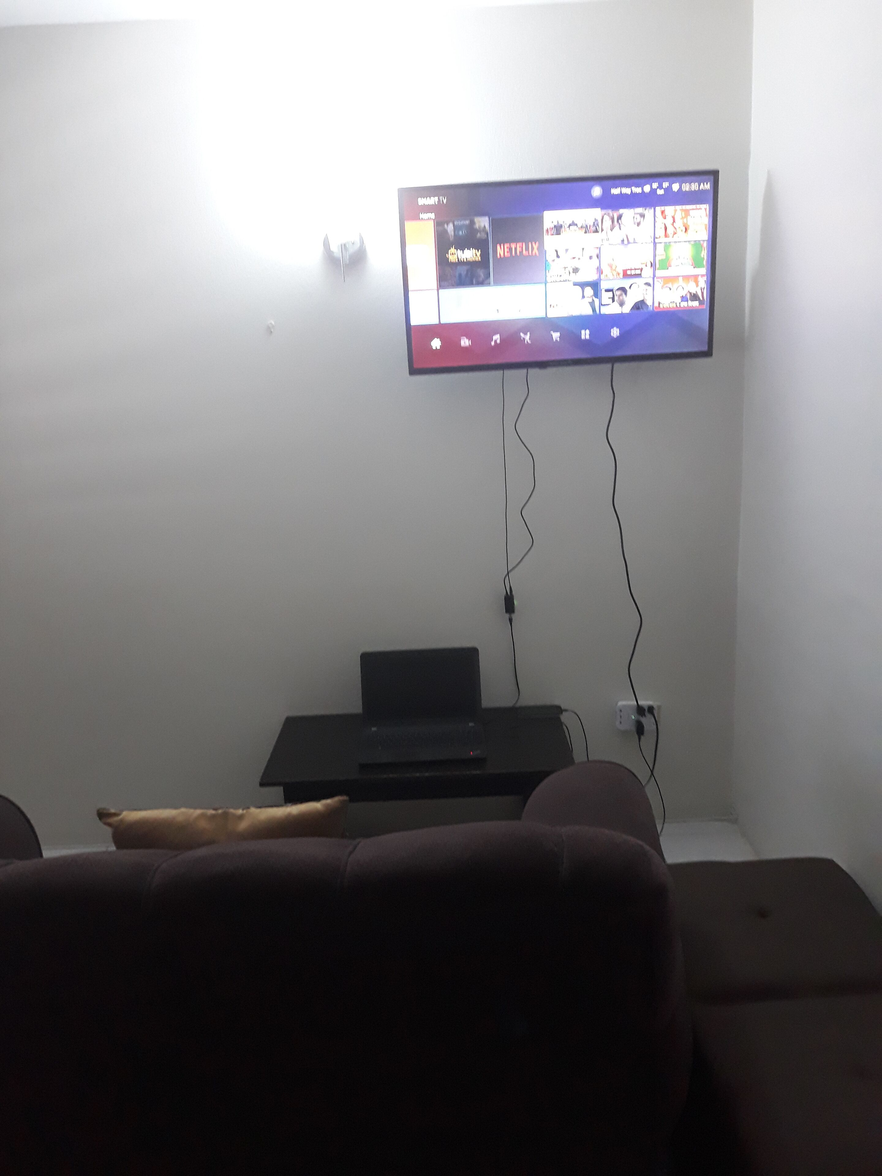 A living room with a mounted TV displaying a streaming service interface, a laptop on a stand, and a couch.