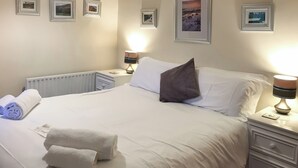 Ein ordentlich bezogenes Bett mit weißen Leinen, einem grauen Kopfkissen und zwei aufgerollten Handtüchern. An der Wand hängen gerahmte Bilder, auf dem Nachttisch steht eine Lampe und es gibt einen weißen Beistelltisch.