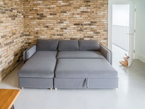 Un canapé d’angle gris est placé contre un mur de briques, avec un balcon à proximité.