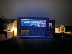Une maison moderne dotée d’une grande fenêtre illuminée, d’un garage et d’une entrée bien éclairée.