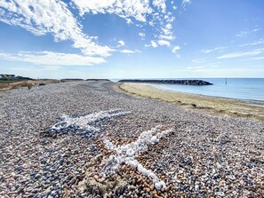 Une plage de galets bordée d’une côte rocheuse et où l’on aperçoit une jetée au loin.