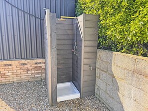Une douche extérieure en bois, dotée d’une structure métallique, d’un mur en briques et d’une haie en arrière-plan.