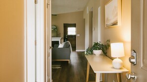 Un couloir avec un plancher en bois, une porte blanche, une petite table avec une lampe, une plante en pot et un tableau encadré au mur.
