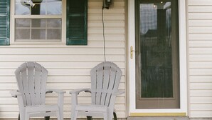 Un porche abrité avec deux fauteuils Adirondack et une petite table, une porte dotée d’un panneau de verre et un luminaire fixé au mur.