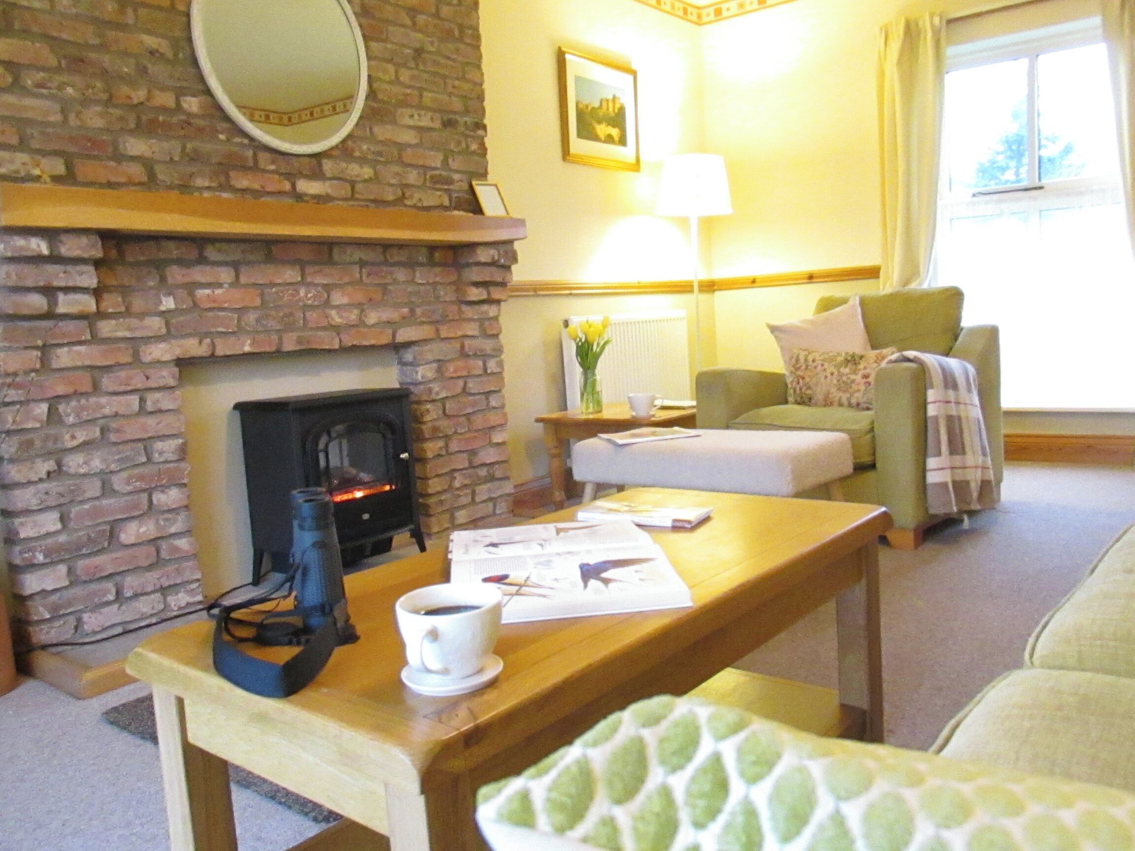 A cozy living room with a brick fireplace, a green armchair, a wooden coffee table, and a lamp.