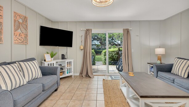 Un salon avec deux canapés gris, un meuble TV blanc, une table basse et une grande fenêtre avec des rideaux.