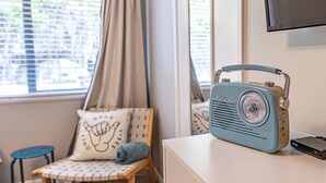 Une pièce avec une vieille radio posée sur une commode, une chaise avec un coussin et une fenêtre avec des stores.