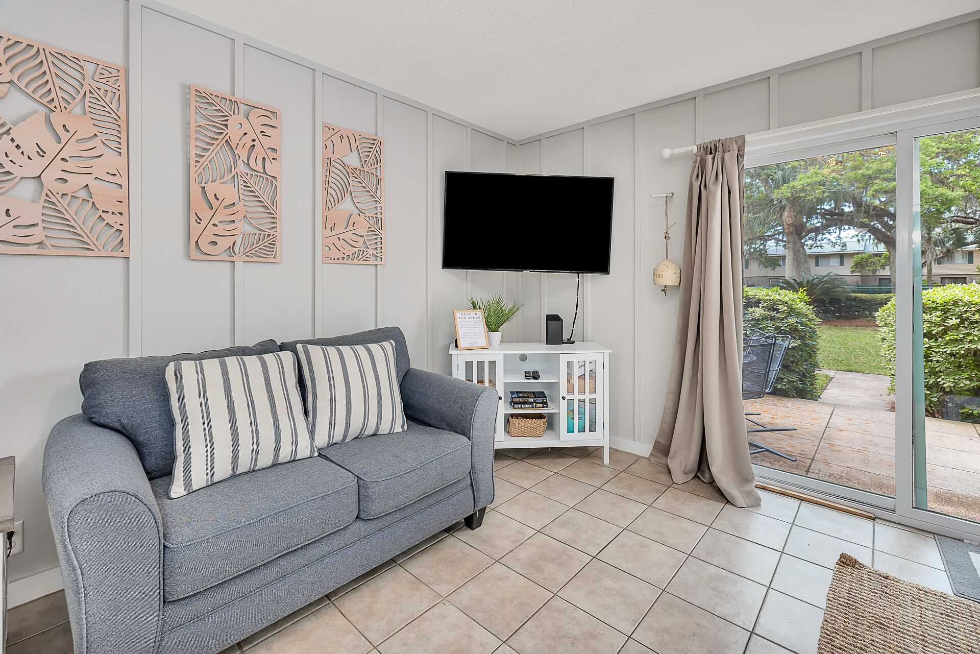 A living room with a grey sofa, a flat-screen TV, a white console table with a plant, and a sliding glass door to a garden.