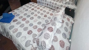 Quarto com duas camas, cada uma com roupa de cama estampada. Há uma janela com cortinas, uma porta e uma pequena mesa com toalha azul.