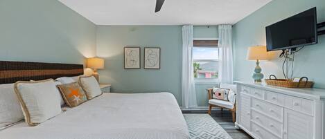 A bedroom with a bed, a ceiling fan, a TV, a nightstand, a lamp, a dresser, and a window with curtains.