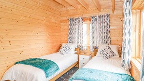 A wooden cabin bedroom with two single beds, a nightstand with a lamp, and patterned curtains. - 2 bedroom accommodation in LLANFAIRPWLLGWYNGYLL (LLANFAIRPWLLGWYNGYLL)
