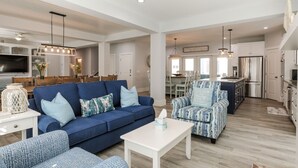 Amplia sala de estar con un sofá azul, un sillón estampado y una mesa de centro blanca. Se ve una cocina con electrodomésticos modernos y un comedor con una araña en el fondo.