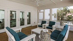 Un área de estar al aire libre techada con mobiliario blanco y cojines azul turquesa, un ventilador de techo y ventanas amplias con vistas a los alrededores.